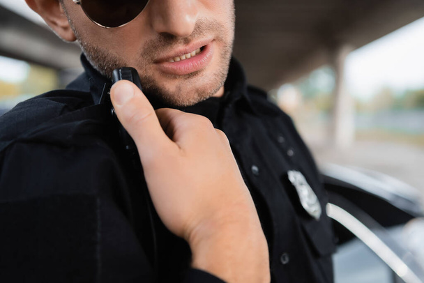 Cropped view of policeman in sunglasses using walkie talkie outdoors  - Фото, изображение