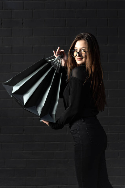 Giovane donna elegante in abiti neri con borse della spesa nere. Muro nero. Telaio verticale. - Foto, immagini