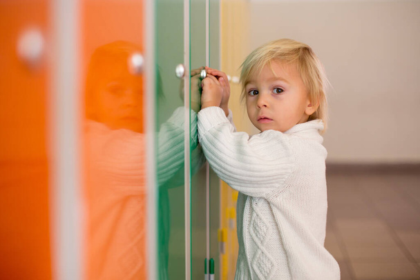 Sweet blonde toddler boy standing in front of a lockers in kindergarden or school hallway, wintertime - Photo, image