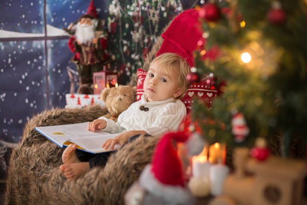 Cute toddler boy, sitting around christmas tree in cozy chair, reading book at christmas night - Фото, изображение