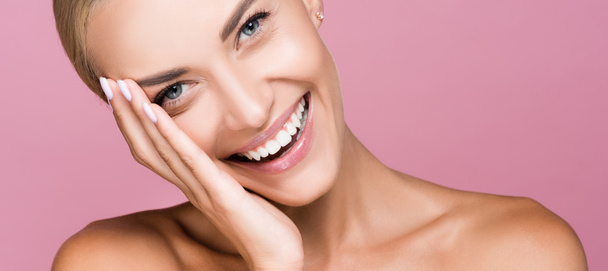 smiling beautiful blonde woman touching face isolated on pink, banner - Photo, Image