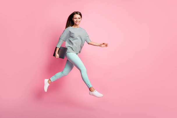 Full size profile photo of cheerful sweet brunette girl run jump hold laptop wear shirt pants sneakers isolated on pink color background - Foto, immagini