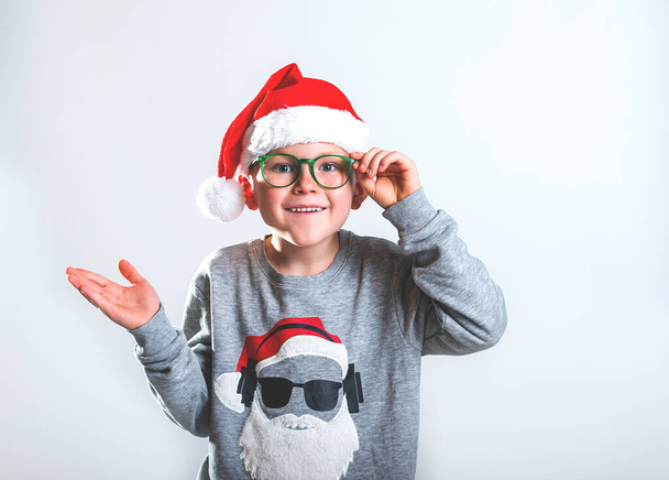 Happy cute boy in red Christmas hat and green glasses. Funny child portrait waiting for Christmas magic. Surprised kid present something with hand with open palm. Stylish school boy isolated on grey - Photo, image