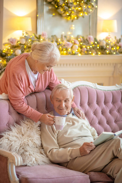 Senior man sitting on a sofa with a book and his wife giving him a cup of tea - Foto, Imagem