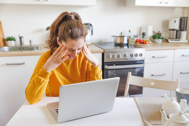 Mobiliroda otthon. Kimerült nő, aki laptopon dolgozik, miközben fej a kézben ül otthon a konyhában. Fiatal lány tanul vagy bent dolgozik. Szabadúszó üzleti karantén koncepció - Fotó, kép