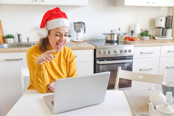 サンタの帽子をかぶっている笑顔の女の子は、ウェブカメラで家族に電話しています。休日に仮想会議チャットを使用して自宅でキッチンに座ってノートパソコンを持つ女性。ハッピークリスマスと新年の新しい通常 - 写真・画像