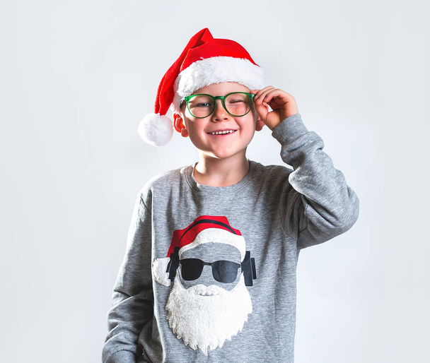 Child boy in christmas red hat and green glasses isolated on grey background. Kid in Santa Claus sweater and funny smiling face winking. Portrait of stylish school pupil at holidays. - Photo, image