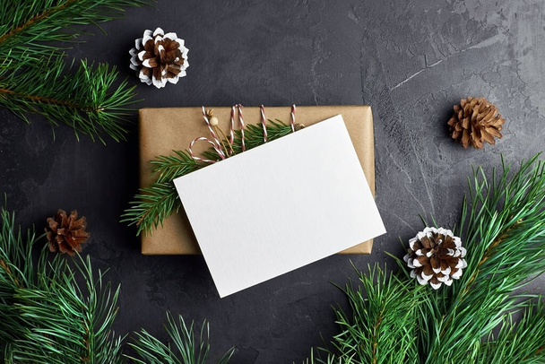 Christmas card mockup with gift box and pine tree branches with cones on dark background. Winter holidays composition, top view, flat lay, copy space. - Photo, Image