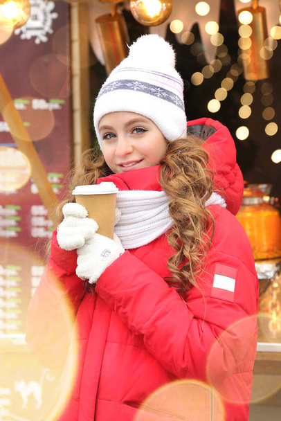 Cute girl in a red long winter coat, a knitted white hat and gloves drinking coffee from a cup.  Boke and flush in the background. Christmas markets. New year concept. Copy space on the right - Photo, image
