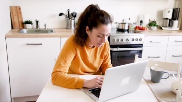 Oficina Móvil en casa. Mujer joven sentada en la cocina en casa trabajando con un ordenador portátil. Chica de estilo de vida estudiando o trabajando en interiores. Concepto de cuarentena empresarial independiente - Metraje, vídeo