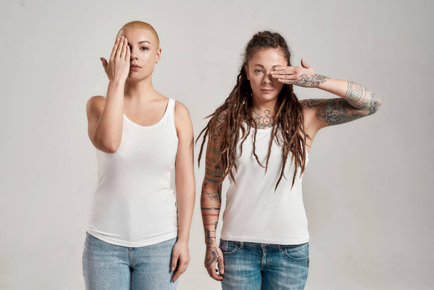Two diverse young caucasian women wearing white shirt, looking at camera, covering half of face with hand while posing together isolated over grey background - Foto, afbeelding