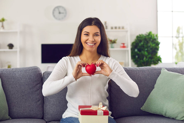 Happy woman sitting on couch with Valentine present on her lap, holding heart and saying thank you - Фото, зображення