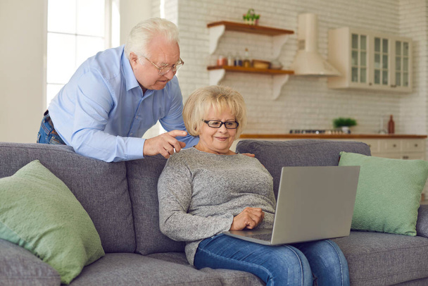 Smiling mature couple staying at home with laptop and communicating or ordering something online - Фото, изображение