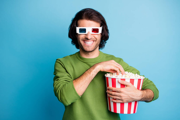 Photo of positive guy hold popcorn box eat shiny smile wear 3d specs green pullover isolated blue color background - Φωτογραφία, εικόνα