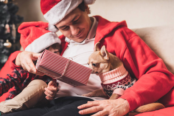 Happy family in Santa hat, father and child son giving Christmas gift at home. Sitting on a couch in the living room with puppy dog in sweater - Foto, Bild