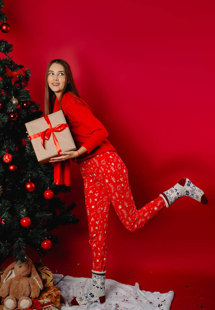 A cute girl next to the Christmas tree holds a gift box, looks away and smiles sweetly. High quality photo - Zdjęcie, obraz