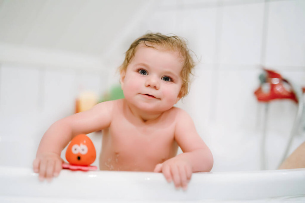 Cute adorable baby girl taking foamy bath in bathtub. Toddler playing with bath rubber toys. Beautiful child having fun with colorful gum toys and foam bubbles - Photo, Image