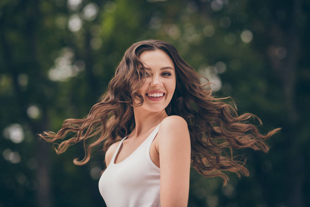 Fénykép édes aranyos fiatal nő mosolygós hosszú göndör barna haj sétáló park viselni fehér felső szabadban - Fotó, kép