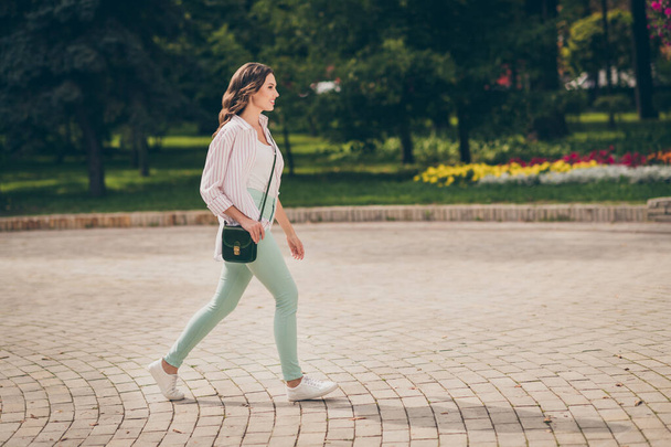 Full size profile photo of cute brunette lady walk park active life wear pastel green jeans white top striped shirt shoulder bag outdoors - Foto, Imagem