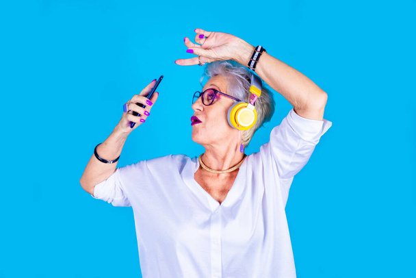 Old woman enthusiastic dancing listening music holding smartphone isolated on blue background enjoying streaming playlist wearing headphones  - Photo, Image
