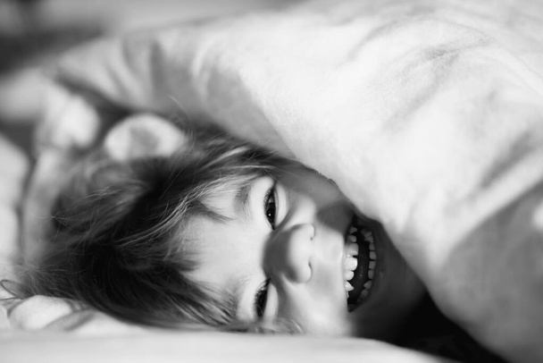 Ένα κοριτσάκι που περνάει καλά στο κρεβάτι μετά το ξύπνημα. Υγιές παιδί άλμα και να παίξετε κρυφτό με τους γονείς. Αξιολάτρευτο παιδί στο σπίτι, οικογενειακή ζωή. - Φωτογραφία, εικόνα