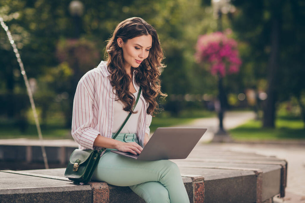 Photo of nice brunette hair young lady sit near fountain work study writing laptop wear green pants shirt top bag in park outdoors - Zdjęcie, obraz