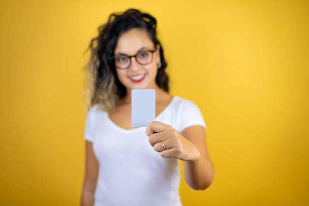 Young beautiful woman wearing casual white t-shirt over isolated yellow background smiling and holding white card - Photo, Image