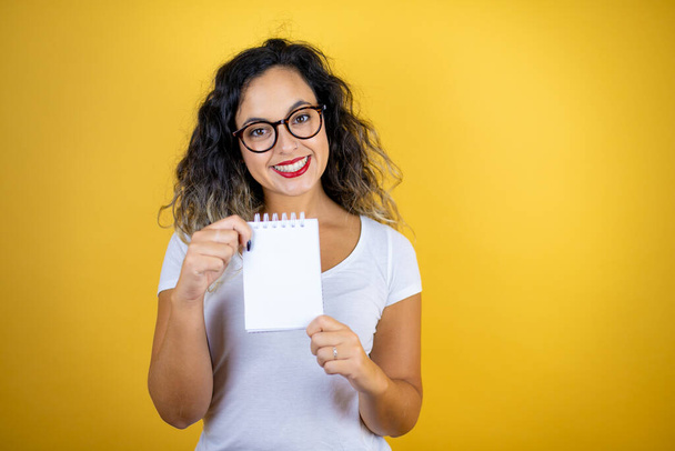 Giovane bella donna indossa casual t-shirt bianca su isolato sfondo giallo sorridente e mostrando notebook vuoto in mano - Foto, immagini
