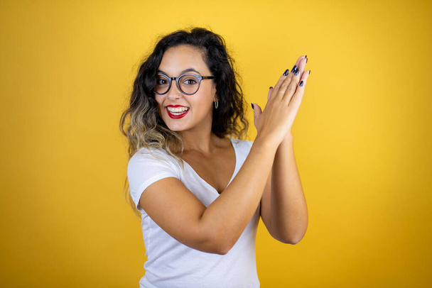 Jonge mooie vrouw draagt casual witte t-shirt over geïsoleerde gele achtergrond klappen en applaudisseren gelukkig en vreugdevolle, lachende trotse handen samen - Foto, afbeelding