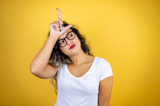 Young beautiful woman wearing casual white t-shirt over isolated yellow background making fun of people with fingers on forehead doing loser gesture mocking and insulting. - Photo, Image