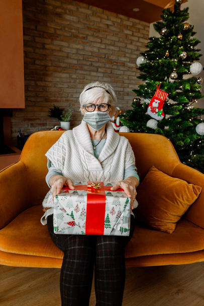 Mujer anciana triste y aburrida con el pelo blanco, sentada sola en casa usando mascarilla. Los no familiares la visitan en Navidad. Autoaislamiento durante la pandemia de Coronavirus. Cuarentena covid, bloqueo. - Foto, imagen