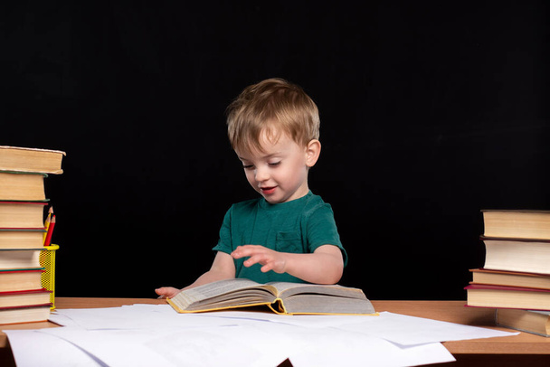 un niño pequeño de aspecto europeo lee un libro en la mesa sobre un fondo negro. Aislar. tarea. - Foto, Imagen