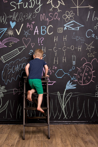 un bambino piccolo di aspetto europeo siede su uno sgabello vicino a una parete di ardesia con disegni. Il ragazzo disegna con un sorriso. home tempo libero - Foto, immagini