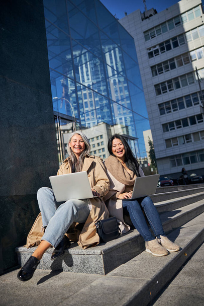 Cheerful female friends with laptops sitting on street steps - Foto, imagen