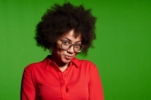 Closeup of shy young African-American girl in glasses and red shirt looking at camera over green background - Foto, imagen