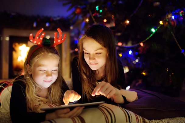Two cute young sisters using a tablet pc at home by a fireplace in warm and cozy living room on Christmas eve. Winter evening at home with family and kids. - Fotó, kép