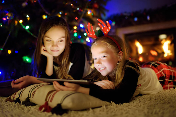 Two cute young sisters using a tablet pc at home by a fireplace in warm and cozy living room on Christmas eve. Winter evening at home with family and kids. - Foto, Imagem