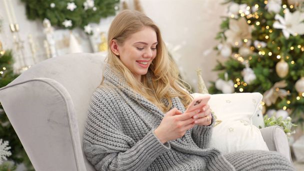 Smiling trendy woman in gray cozy sweater sittin on comfortable armchair and sending text message - Photo, Image