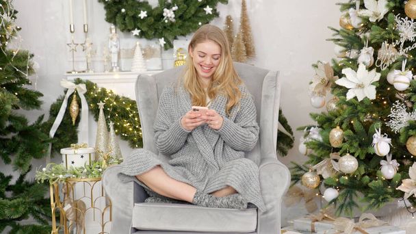 Donna sorridente alla moda in maglione grigio accogliente seduto su una comoda poltrona e l'invio di messaggi di testo - Foto, immagini