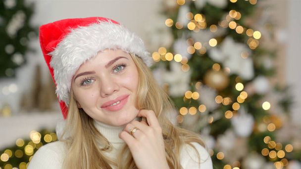 Young blond beautiful smiling woman in sweater and Santa hat on christmas tree background - Foto, Imagem