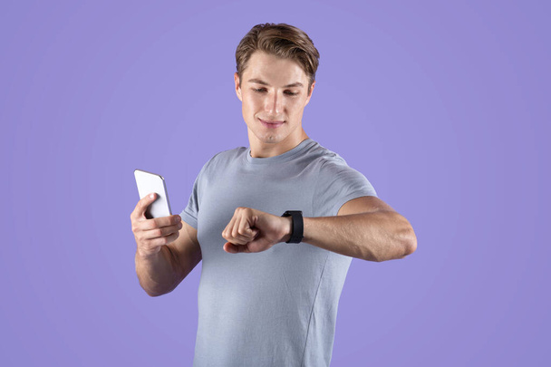 Active millennial guy with modern smartwatch and cellphone over violet studio background - Photo, image