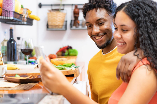 幸せな若いカップルは、タブレット上でレシピを見て。美しい若いカップルは、自宅でキッチンで健康的な食べ物を準備笑顔。テクノロジー、愛、食の概念. - 写真・画像
