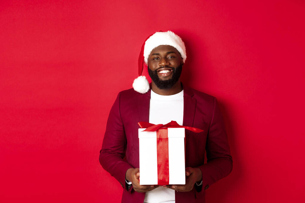 Kerstmis, Nieuwjaar en winkelen concept. Gelukkig bebaarde Afro-Amerikaanse man met xmas aanwezig, glimlachend op camera, staande in santa hoed tegen rode achtergrond - Foto, afbeelding