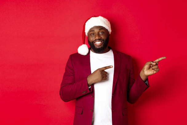 Karácsony, parti és ünnepek koncepció. Mosolygó fekete férfi szakállal és télapó kalap, ujjal mutogatva a logó, felett áll piros háttér - Fotó, kép