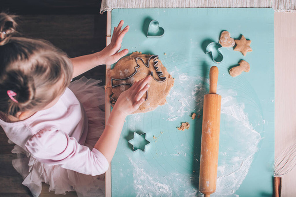 Милая очаровательная малышка режет печенье из свежего теста и печет пряничное рождественское печенье на кухне. - Фото, изображение