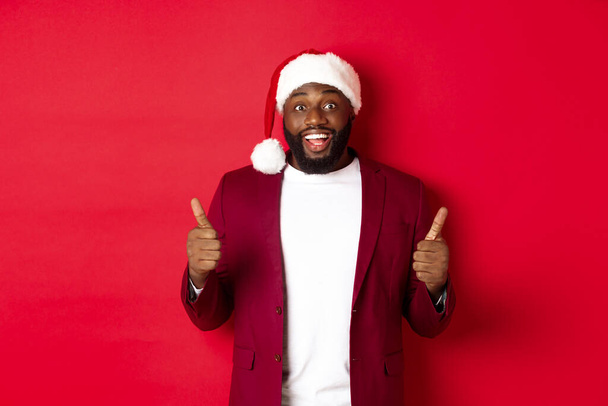 Christmas, party and holidays concept. Cheerful Black man in santa hat showing thumbs-up, celebrating New Year, approve and agree, standing against red background - Zdjęcie, obraz