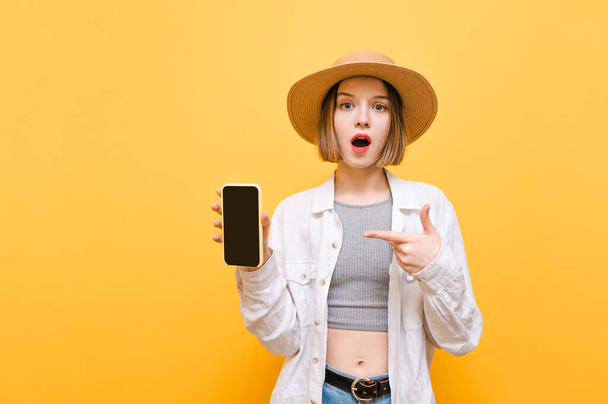 Portrait of surprised lady in summer clothes and hat stands on a yellow background, points her finger at the black screen of a smartphone in her hand and looks into the camera with a shocked face - Photo, Image