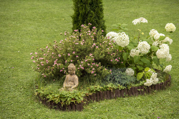 Meditierende Buddha-Figur sitzt in einer kleinen Blumeninsel im Blumenbeet, umgeben von natürlicher Holzpalisade. - Foto, Bild