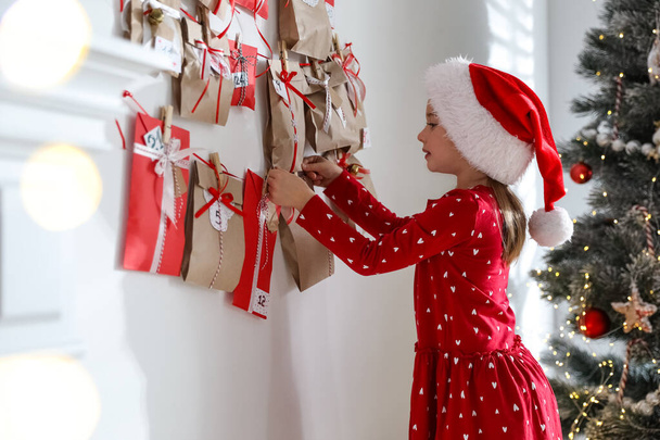 Cute little girl in Santa hat taking gift from Christmas advent calendar at home - Zdjęcie, obraz