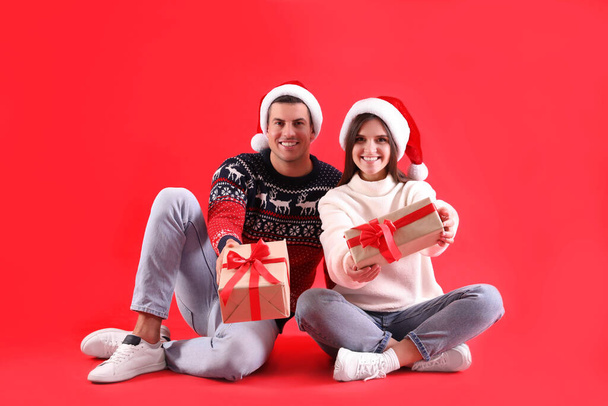 Красивая счастливая пара в шляпах и свитерах Санты сидя с рождественскими подарками на красном фоне - Фото, изображение